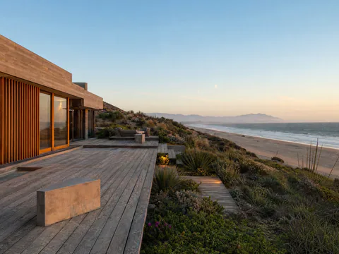 Casa Duna: volúmenes de concreto cimbrado y celosía de madera, con una terraza de madera que baja entre las dunas a la playa del Pacífico, al atardecer