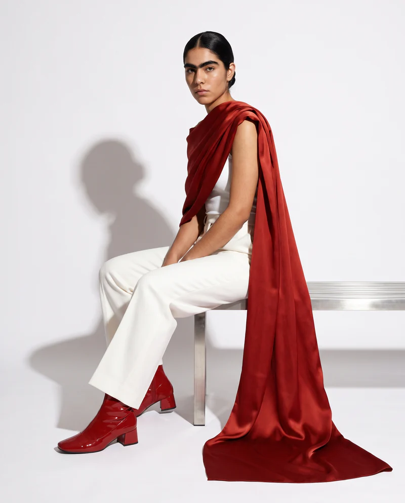 La modelo sentada en una banca de acero, pantalón blanco de tiro alto y estola de seda roja hasta el piso